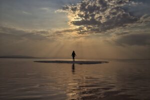 A person standing on a very small island surrounded by water, the sun filtering through grey clouds, a scene of quiet reflection