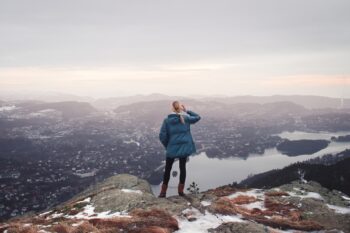 A person standing on a snowy peak, overlooking the countryside and lake below. They've reached their highest point. What's your mountain to climb?