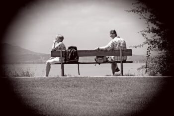 Two people sat on a bench. One is looking at something, one is looking away. What are they communicating to you and to each other?