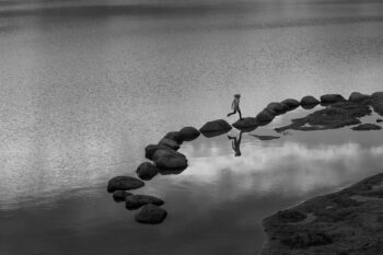 A person in a black and white photo, jumping from rock to rock on the edge of a lake. It's all about having faith in your ability to succeed.