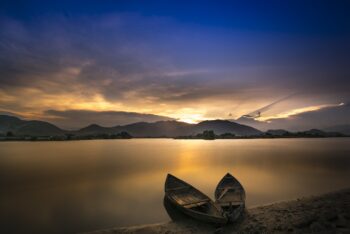 Boats resting on the shore of a completely calm lack, hills in the distance as the sun sets.